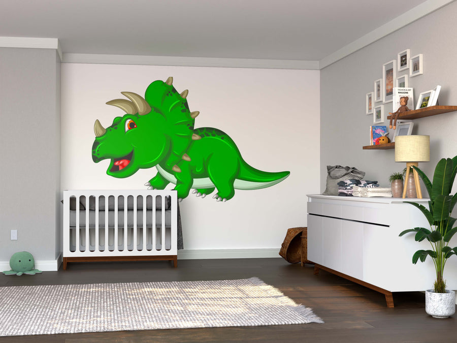 A nursery with wood floors, a white crib, and the Happy Triceratops Wall Mural. A dresser holds a plant and framed photos; shelves display toys and books. A stuffed turtle sits on a round rug near the crib.