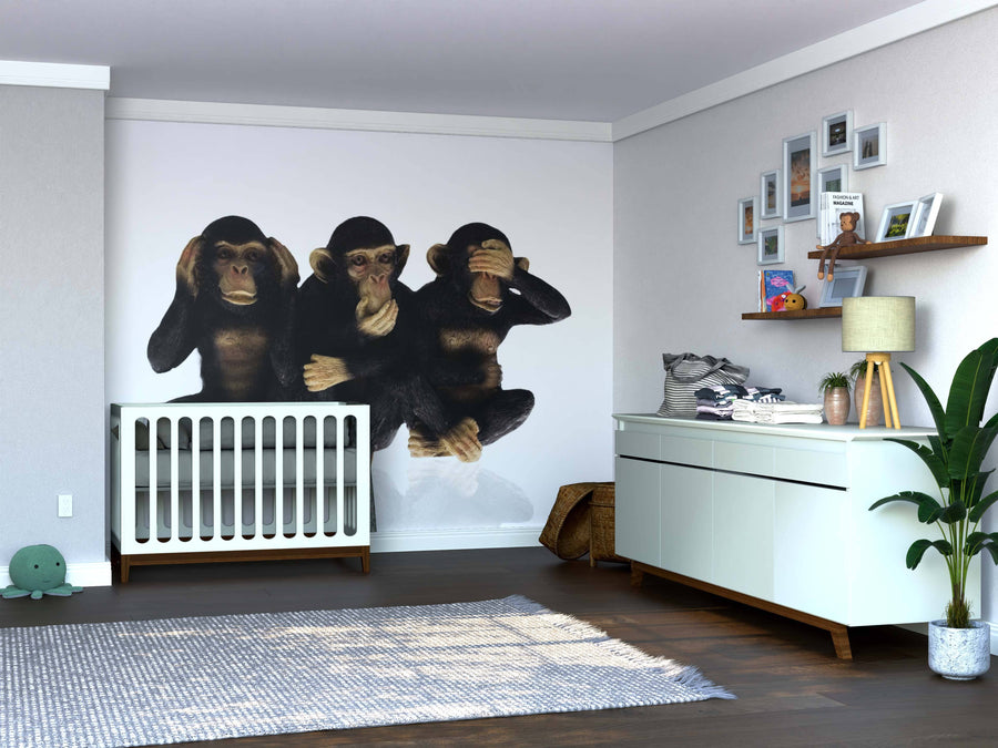 A nursery with wooden floors features a crib against the Hear Speak See Wall Mural, showing three monkeys. To the right is a white dresser with baby items and a shelf holding framed photos, while a rug and plant complete the decor.