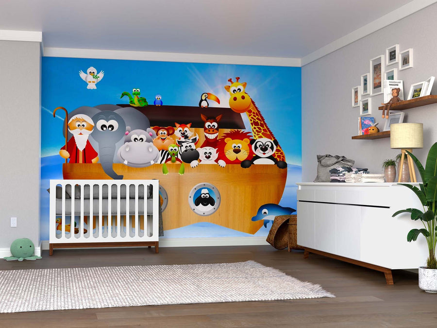 A nursery with a white crib, dresser, plant, and toys. The wall features the Joyful Voyage Wall Mural with cartoon animals and Noah under a blue sky. Wooden floors and soft decor complete the look.