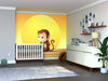 A nursery with a crib in front of the Monkey Reading Wall Mural showing a smiling cartoon monkey and yellow sun, plus a white dresser, potted plant, wall shelves with decor, framed photos, and a soft toy turtle on the floor.