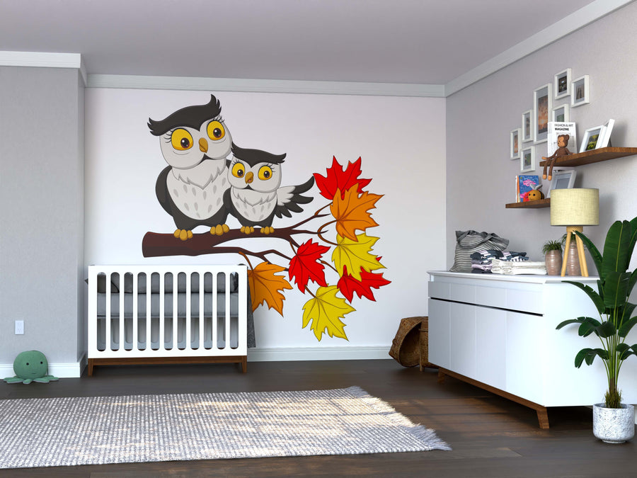 A nursery with gray walls and a white crib features the Mother and Baby Owl Wall Mural—a peel-and-stick design of two cartoon owls on a branch with autumn leaves. A dresser, plant, rug, framed photos, and toys complete the cozy space.