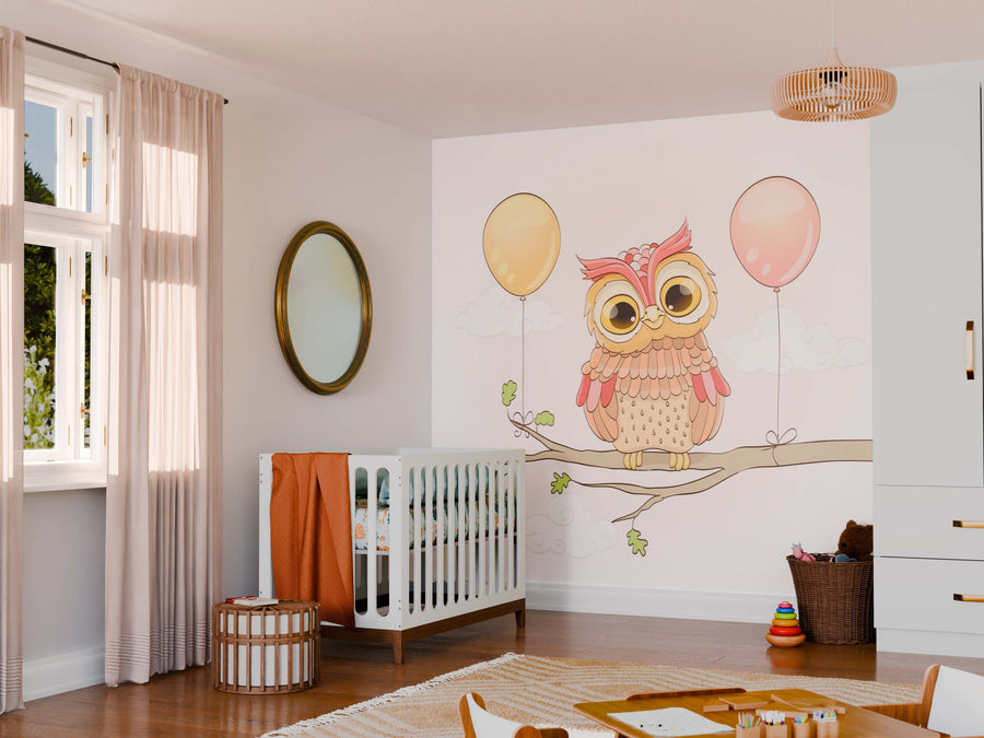 A cozy nursery with light walls features a crib, round mirror, woven storage, toys, and the Owl with Balloons Wall Mural depicting a cute cartoon owl on a branch. Soft sunlight filters through sheer curtains on the window.