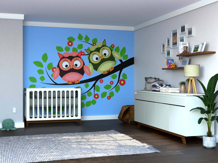 A nursery with light walls, a white crib, and the Owl Duo Wall Mural showing two cartoon owls on a leafy branch. Features include a dresser with decor, floating shelves with frames, potted plant, gray rug, and wooden floors for playfulness.