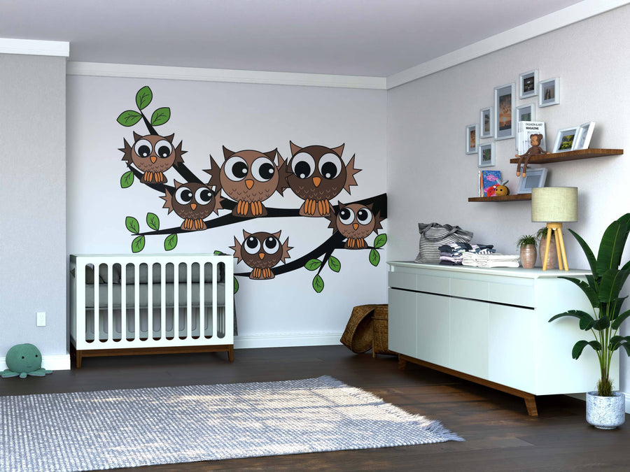 A modern nursery with a white crib, dresser, plant, and shelves features the Owl Family Wall Mural of five cartoon owls on branches. Wooden floors, neutral walls, and playful decor create a cozy atmosphere.