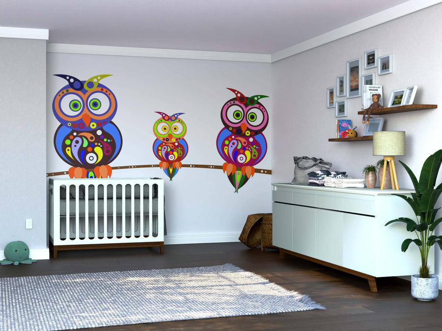 A nursery room features a white crib, the Owl Kinship Wall Mural with colorful cartoon owls, a dresser with a plant, a basket, and shelves displaying decor—all on a dark wood floor and light-colored walls.