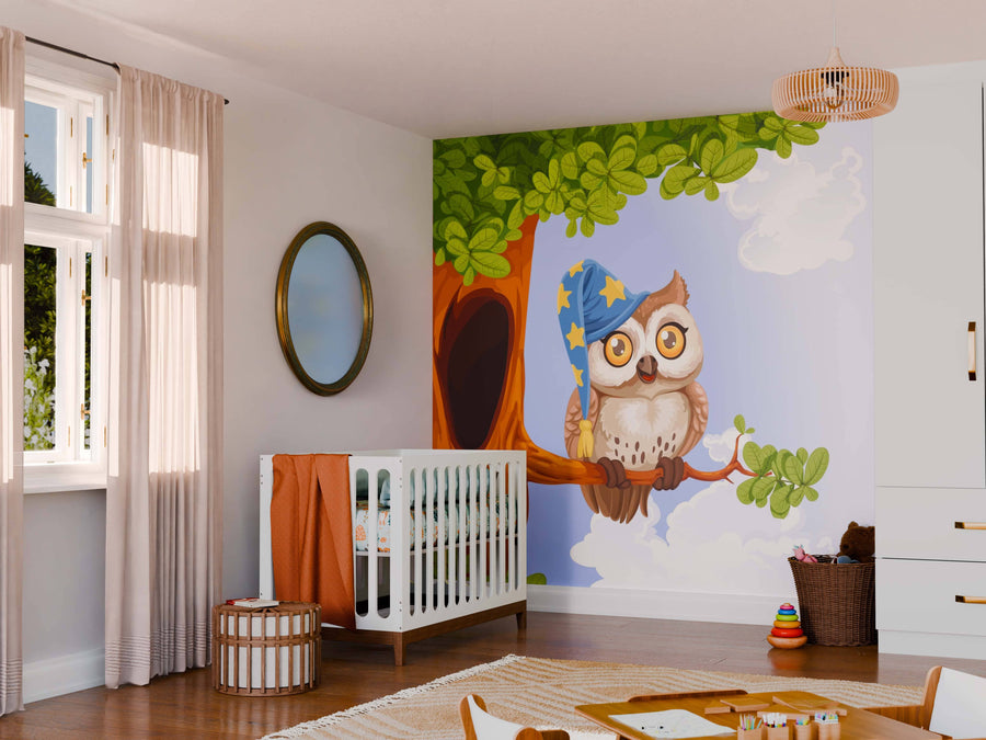 A nursery with a crib, round mirror, and toys. The wall features the Owl Wearing Hat Wall Mural, showing an owl in a wizard hat on a tree branch with green leaves and blue sky. Warm sunlight comes through curtained windows.