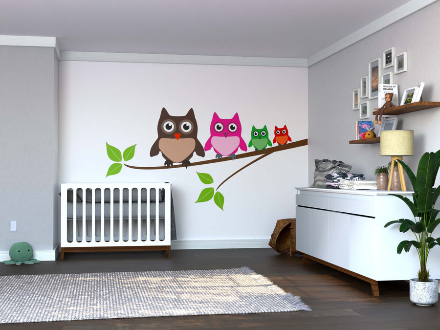 A modern nursery with a white crib, dresser, and plant features the Owls Wall Mural—four colorful cartoon owls on a branch—adding playful charm above wooden flooring and framed pictures on the shelf.