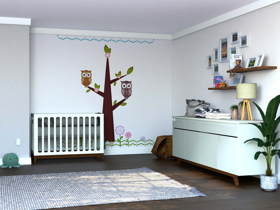A modern nursery features a crib, dresser, and the Owls on Branches Wall Mural. Family photos, a potted plant, rug, and turtle plush toy add charm to the softly lit wooden floor.