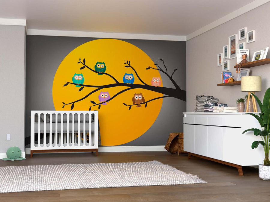 A nursery with a crib, dresser, plants, and shelves features the Owls Under Full Moon Wall Mural—a large yellow peel-and-stick circle with a tree branch and six colorful cartoon owls. Light walls, wood floors, and photos decorate the space above the dresser.