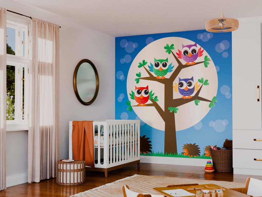 A nursery with a white crib, wicker storage, and neutral decor features the Owls at Night Wall Mural—peel and stick art of cartoon owls in a moonlit tree over hedgehogs—while sunlight filters through sheer curtain panels.