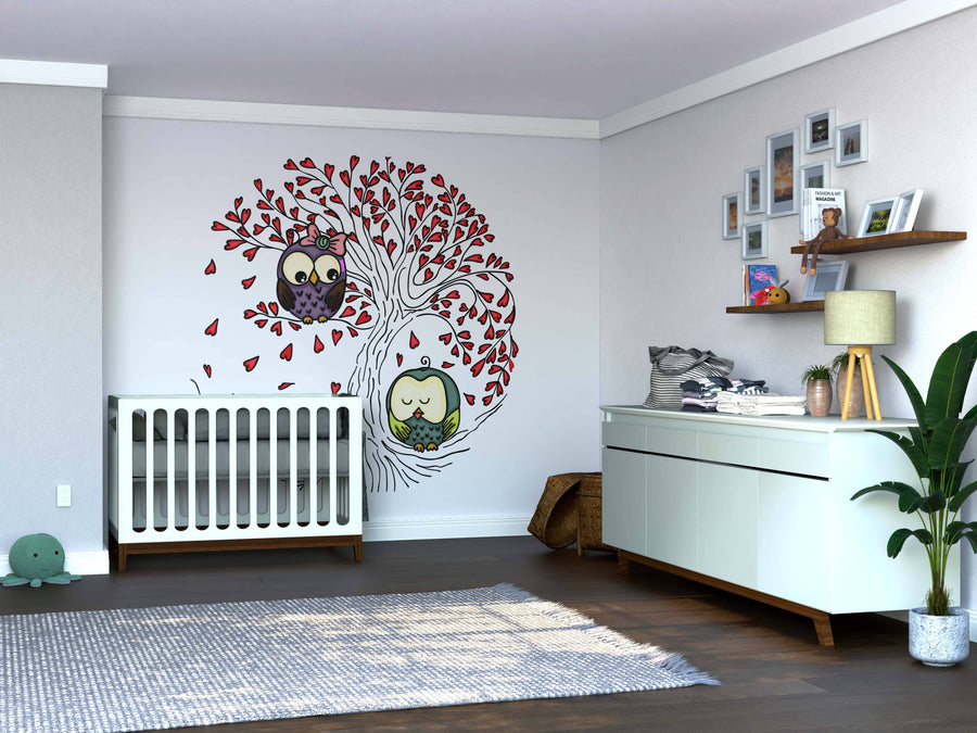 A modern nursery features a white crib next to the Owls in Love Tree Wall Mural, showing two cartoon owls in a tree with red leaves. A white dresser, plant, shelf with framed photos, and a gray rug complete the decor on wooden floors.