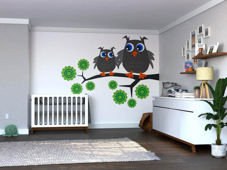 A modern nursery with a white crib, dresser, and plant features the Pair of Gray Owls Wall Mural—two cartoon owls on a branch with green flowers. Shelves and framed pictures hang above the dresser; a gray rug and green turtle plush sit on the floor.