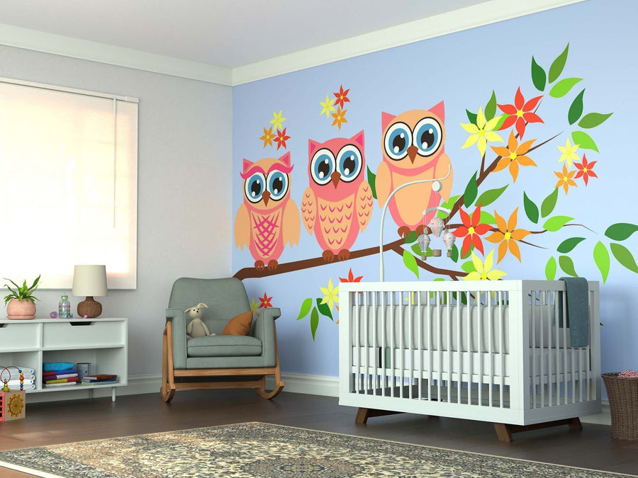 A bright nursery with a crib, armchair, and shelf features wooden floors, a patterned rug, and the Pink Owls Wall Mural—a large custom print of three colorful cartoon owls with big eyes perched on a leafy branch.