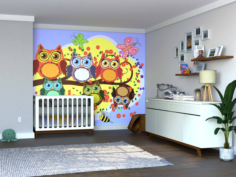 A bright nursery features the Playful Forest Scene Wall Mural, displaying colorful cartoon owls, butterflies, and flowers on a tree. A white crib is set against it, with a dresser, framed photos, a potted plant, and a rug completing the cozy decor.