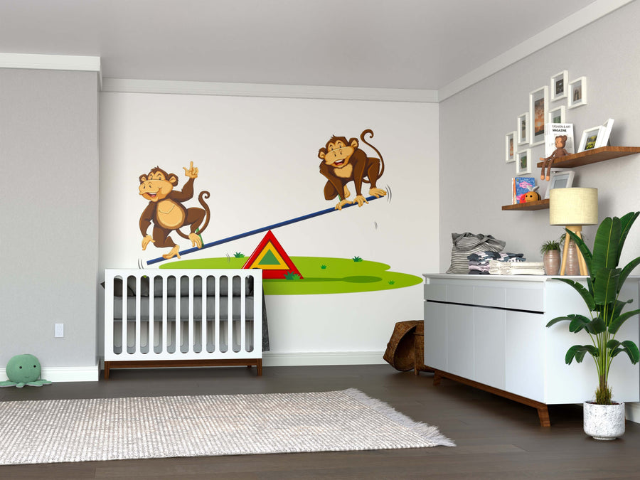 A nursery room with a white crib and dresser features the Playful Monkey See-Saw Wall Mural, showing two cartoon monkeys on a seesaw over green grass. A rug, toys, and shelves with books and decor complete the cheerful space.