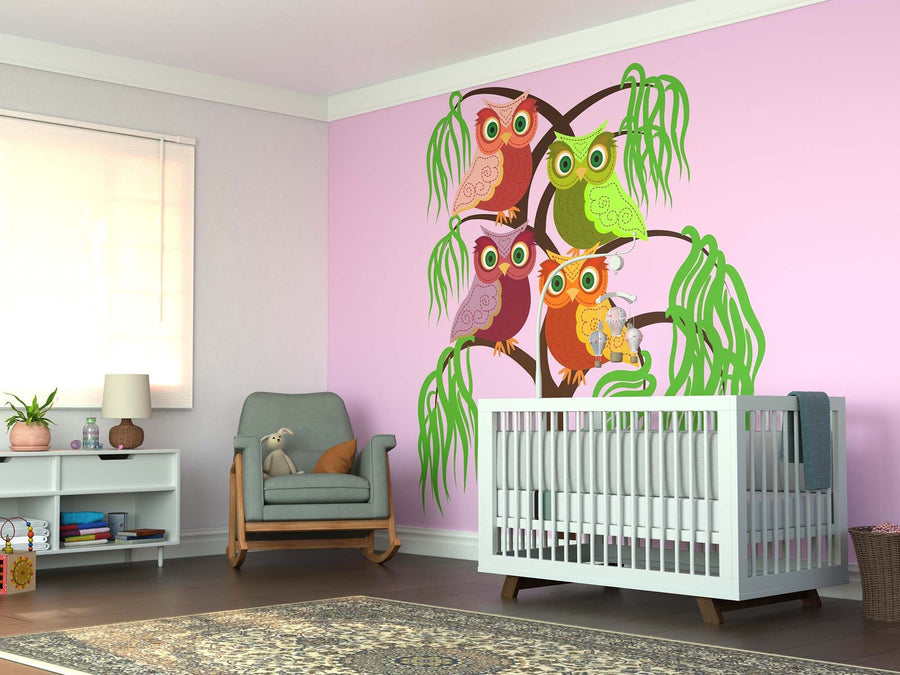 A nursery with pink walls showcases the Playful Owls Wall Mural—four vibrant owls on a tree. The room includes a white crib, green armchair with a stuffed rabbit, toy shelves, and a patterned rug over the wooden floor.