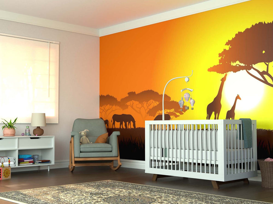 A nursery room features a white crib, gray armchair, shelves, and the Savanna Sunset Wall Mural—a peel and stick mural showing giraffe and elephant silhouettes under a yellow-orange sunset. A rug and baby mobile complete the cozy space.