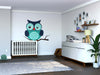 A modern nursery with a white crib, dresser, plant, and shelves with photos and toys. The Sleepy Blue Owl Wall Mural adds playful teal and blue charm. Wood floors and neutral walls complete the cozy vibe.