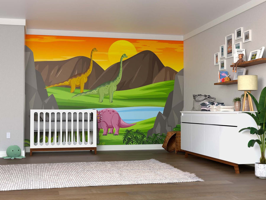 A nursery with a crib, white dresser, plants, toys, and shelves has its back wall decorated with the Sunset in Dino Valley Wall Mural—featuring mountains, a river, and three cartoon dinosaurs beneath a yellow-orange sunset sky.
