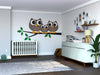 A modern nursery with a white crib and dresser, plant, toys, floating shelves for photos and books, wood floors, neutral walls, and the Sweet Owl Family Wall Mural featuring three cartoon owls on a branch above the crib.
