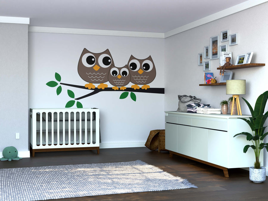 A modern nursery with a white crib and dresser, plant, toys, floating shelves for photos and books, wood floors, neutral walls, and the Sweet Owl Family Wall Mural featuring three cartoon owls on a branch above the crib.