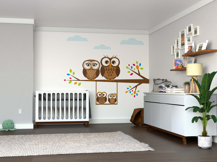 A modern nursery with a white crib and dresser features the Swinging Owl Family Wall Mural—peel and stick accents in neutral tones with colorful owls on branches—plus baby supplies and a large green plant.