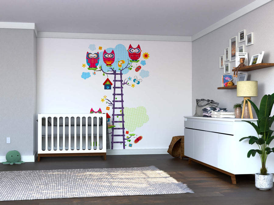 A modern nursery featuring a white crib, baby dresser, green plant, and an Owl Tree Wall Mural with colorful owls. The room has hardwood floors, gray walls, framed photos on floating shelves, a ladder, and playful cloud accents.