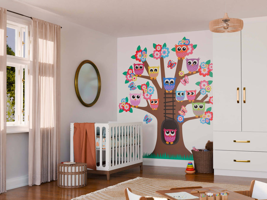 A nursery features a crib, storage baskets, and the Tree of Owls Wall Mural—a large, colorful peel-and-stick design with cartoon owls and flowers. Soft light through beige curtains and a white wardrobe complete the cozy, playful room.