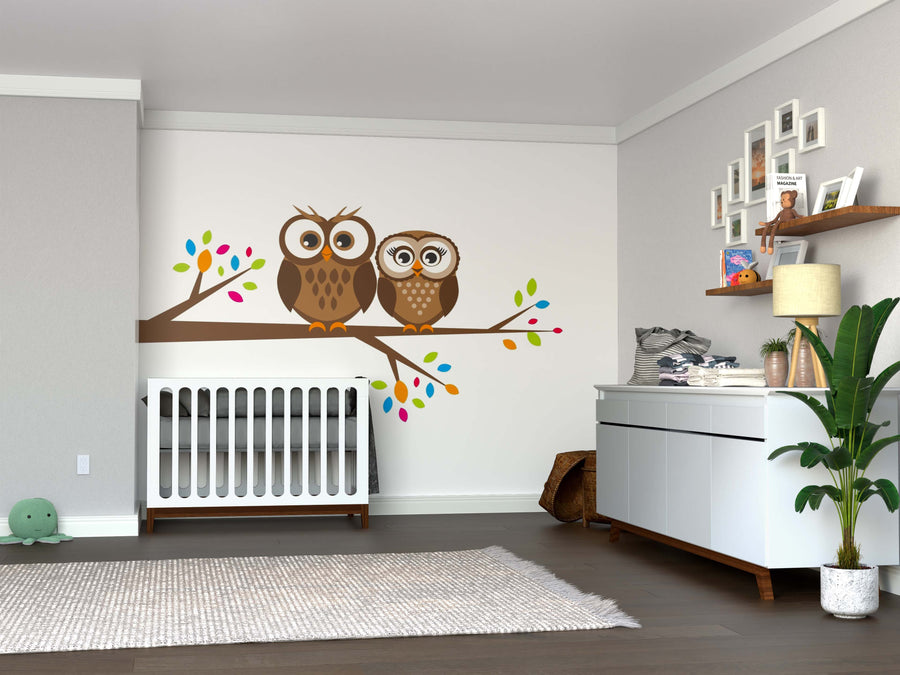 Modern nursery with a white crib, light gray walls, the Two Brown Owls Wall Mural featuring two cartoon owls on a leafy branch, white dresser, shelves with books and decor, a plant, and a gray rug on wooden floor.