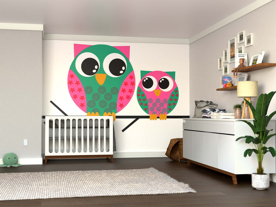 A nursery with wooden floors and neutral tones is playfully decorated with a crib, dresser, toys, plants, and the Two Colorful Owls Wall Mural featuring a large and small cartoon owl on a branch.
