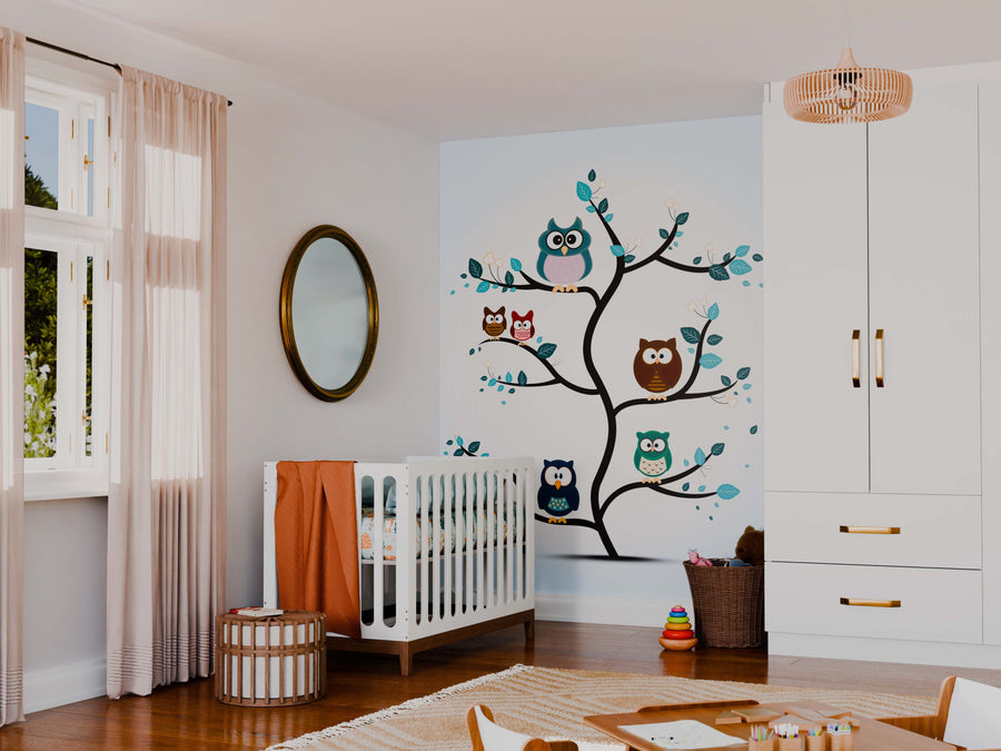 A cozy nursery with a white crib, orange blanket, soft rug, wicker basket, toy stackers, sheer curtains, and rattan light fixture. The wall features the Whimsical Owl Family Wall Mural with five cartoon owls in a tree.