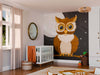 A cozy nursery features the Wise Moon Watcher Wall Mural—a custom peel and stick mural showing a wide-eyed cartoon brown owl against a nighttime sky—along with a crib, toys, soft decor, beige curtains, and a round mirror.