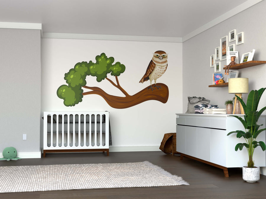A nursery with gray walls features a white crib under the Wise Tree Watcher Wall Mural, showing an owl on a branch. To the right, there’s a white changing table with decor above, a plant beside it, and a soft gray rug on the wooden floor.