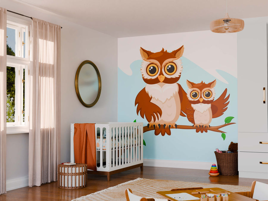 A cozy nursery features the Wise and Watchful Wall Mural of two cartoon owls on a branch, complemented by a white crib with orange bedding, a round mirror, woven basket, toy stack, soft curtains, and light wood furniture in the sunlit room.