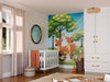 A bright nursery features a crib, round mirror, changing table, and the Woodland Friends Wall Mural with cartoon deer, rabbits, and trees under a blue sky. Sunlight shines through sheer curtains onto neatly arranged toys and baskets.