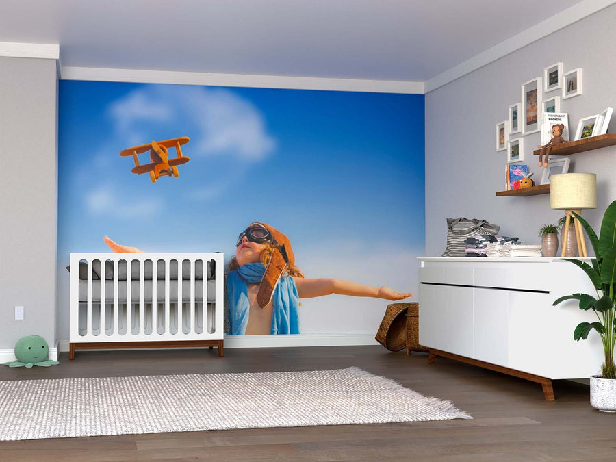 A modern nursery with a white crib, dresser, and plants features the Young Aviator Wall Mural—showing a child in aviator goggles playing with a toy plane against a blue sky. Shelves display toys and framed photos.