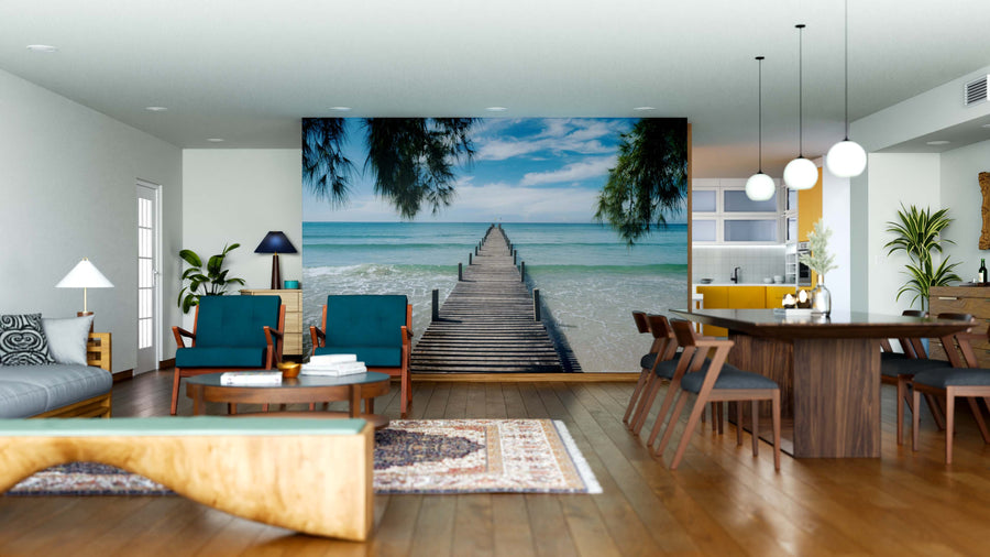 A modern living-dining room boasts wood floors, blue and wood furniture, pendant lights, and an Ocean Pathway Wall Mural depicting a wooden pier leading into a tropical ocean beneath a blue sky. Natural light enters from the left window.