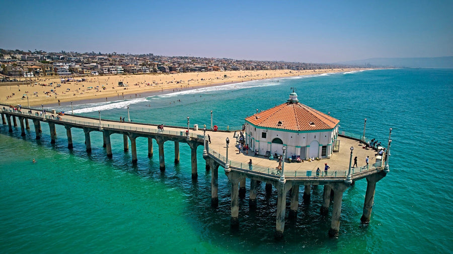 A modern living-dining room with wood furniture and rugs features the Ocean View from Pier Wall Mural, showing a seaside pier with a round building over turquoise water. The kitchen's yellow accents can be seen beyond the mural.