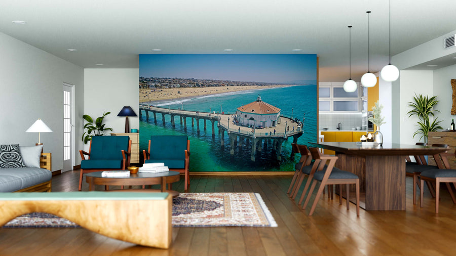 A modern living-dining room with wood furniture and rugs features the Ocean View from Pier Wall Mural, showing a seaside pier with a round building over turquoise water. The kitchen's yellow accents can be seen beyond the mural.