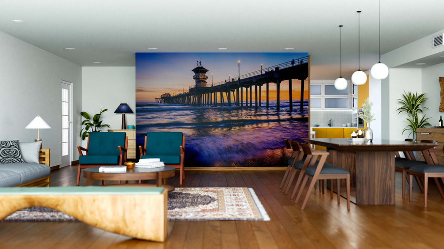 Modern living-dining space with wooden floors and blue-green chairs features the Ocean and Pier Wall Mural—a custom print of a sunset pier over ocean waves. Pendant lights hang above a dark dining table, near a beige sofa and patterned rug.