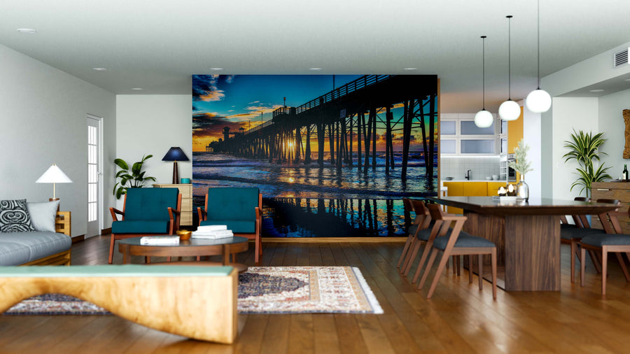 A modern living and dining area with wood floors, dark blue and wood furniture, and the Oceanside Pier at Sunset Wall Mural. Spherical pendant lights above the dining table highlight vivid orange and blue tones from the mural.