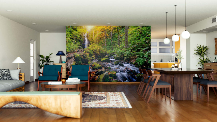 A modern, open-plan living and dining area features the Oirase Autumn Fall Landscape Wall Mural—a lush forest with a stream and sunlight. Wooden furniture, teal chairs, a rug, and a minimalist kitchen create a cozy, nature-inspired vibe.