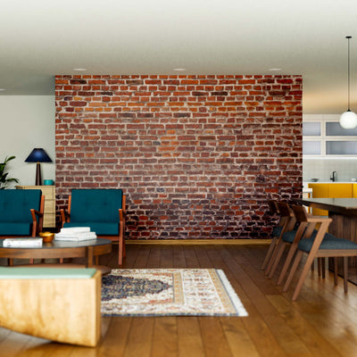 Modern open-plan living and dining area with an Old Brick Facade Wall Mural, wooden floors, dark green chairs, gray sofa, large dining table with chairs, pendant lights, indoor plants, and a kitchen with yellow cabinets in the background.
