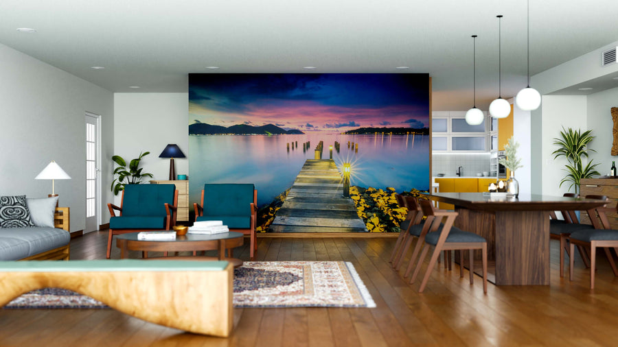 A modern living and dining room features wooden furniture, blue chairs, and the Old Bridge Wall Mural depicting a dock over calm water at sunset. Pendant lights, a kitchen area, and a carpet on the wood floor add to the cozy atmosphere.