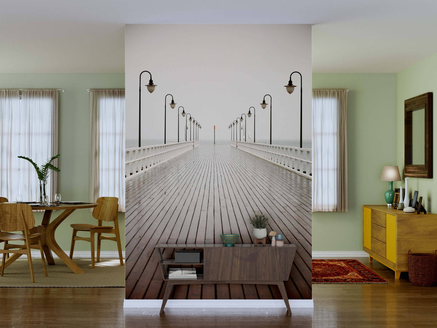 A bright living area with dining space and yellow sideboard is divided by the Old Pier Wall Mural, depicting a wooden pier with black lamp posts in misty water; a modern console with plants and decor stands in front.