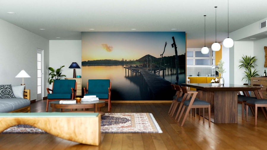 Modern open-plan living and dining room with wooden floors, blue chairs, and the Overwater Oasis Wall Mural—a peel and stick mural of a lakeside pier at sunset. Pendant lights, plants, a rug, dining table, and minimalist decor complete the sunlit space.