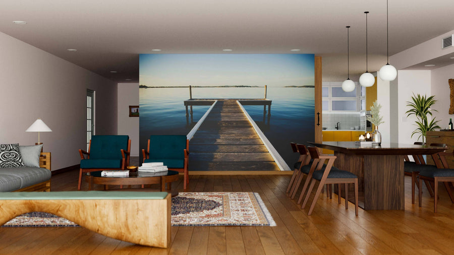 A modern living-dining room features wood furniture, green chairs, and a large Paradise Pier Wall Mural over calm water. Pendant lights hang above the dining table, filling the open space with warm lighting and neutral walls.