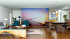 A modern living-dining space with wooden floors, green chairs, pendant lights, indoor plants, and the Pastel Pier Sunset Wall Mural creates a bright, airy contemporary vibe. The kitchen is visible for an open feel.