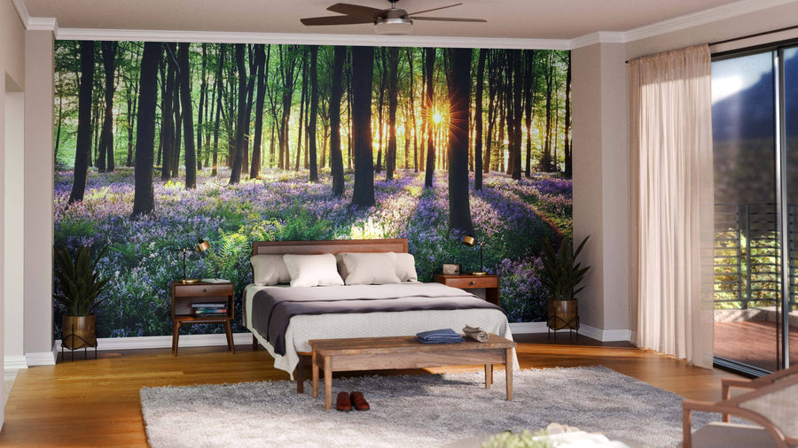 A modern bedroom features the Path Through Bluebell Woods Wall Mural, showcasing a sunlit forest with tall trees and purple wildflowers. The room includes a bed with neutral bedding, wooden nightstands, a rug, bench, ceiling fan, and large glass doors.