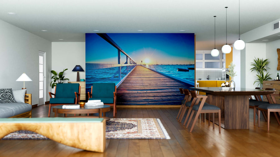 Modern open living and dining area with wooden floors, teal chairs, and the Path to Sea Wall Mural—a stunning peel and stick boardwalk sunset. Pendant lights hang over a dark table; a bright kitchen with yellow accents is in the background.